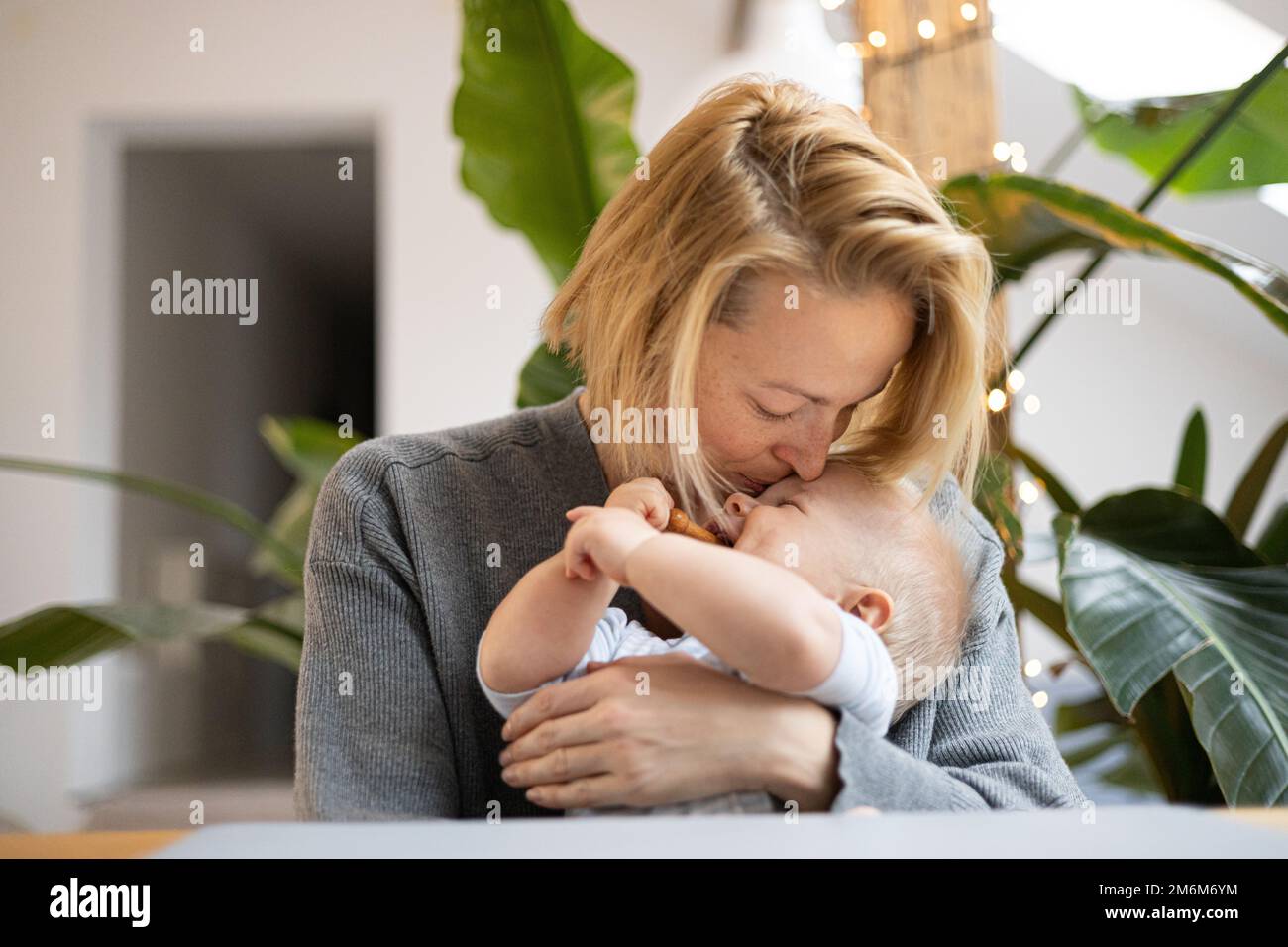 Porträt einer jungen Mutter, die ihren niedlichen kleinen Jungen kuschelt und küsst, während sie zu Hause am Tisch sitzt. Sensorische Stimulation für Babyentwickler Stockfoto
