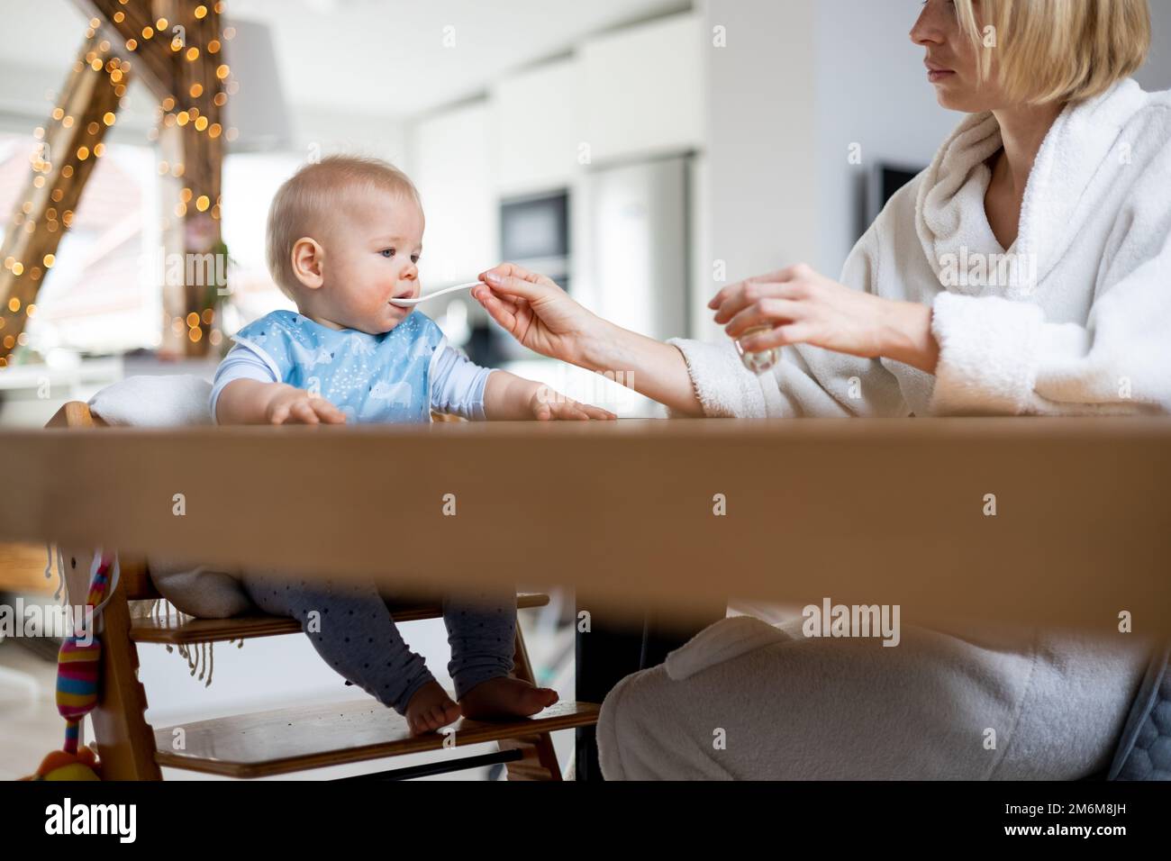Mutter trägt einen gemütlichen Bademantel Löffel und füttert ihr Kind im Baby-Hochstuhl mit Fruchtpüree am Esstisch zu Hause. Einführschleuse für feste Babynahrung Stockfoto