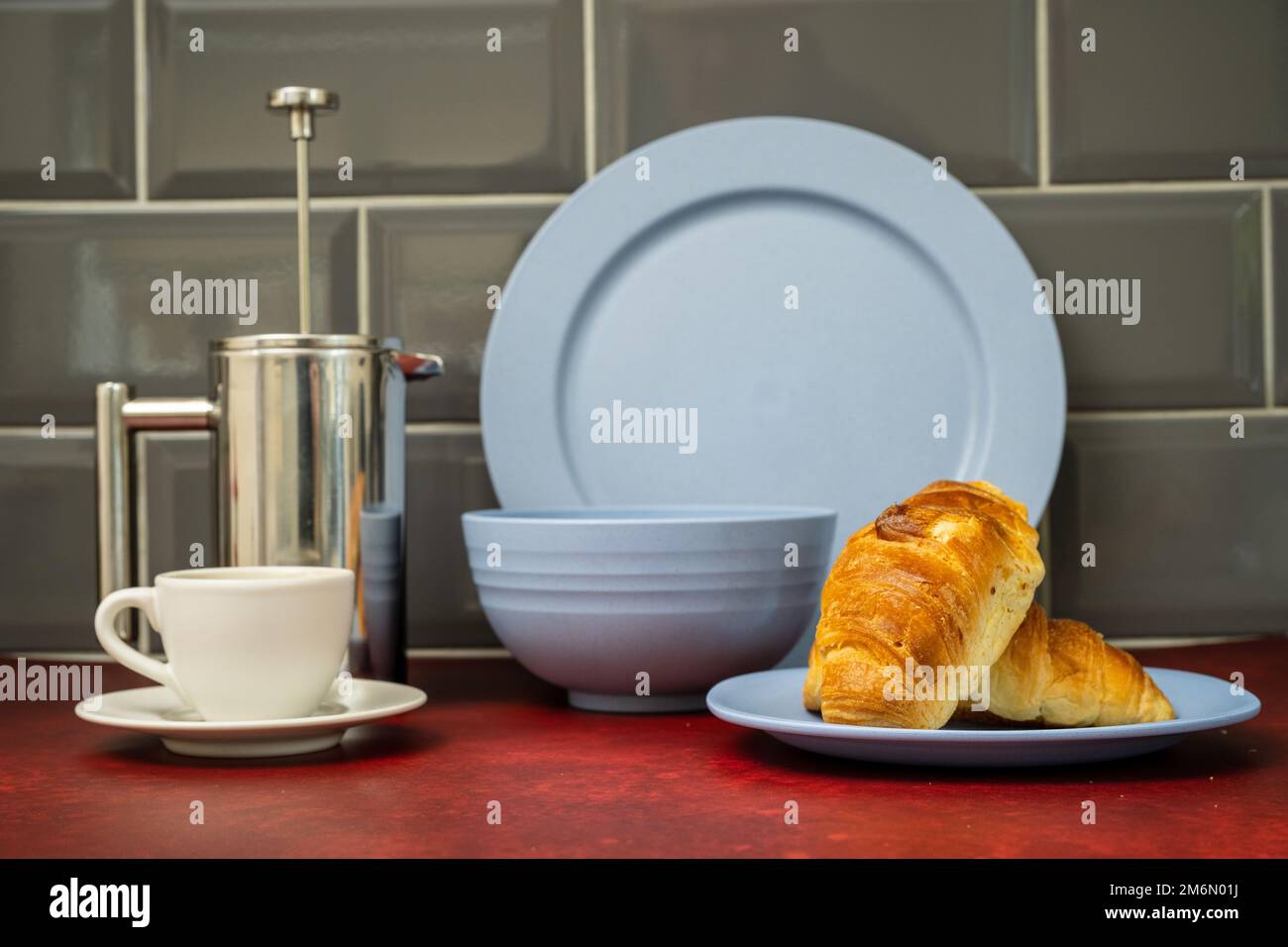 Espresso-Kaffeetasse und Untertasse aus Edelstahl cafetiere mit frischem Coissant Blue Dinner-Set auf rotem Linoleum Arbeitsplatte mit grauem Fliesen auf der Rückseite Stockfoto
