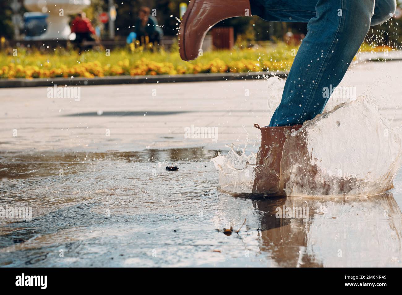 Frau mit Regengummistiefeln, die beim Herbstregen mit Wasserspritzern und Tropfen in die Pfütze laufen und springen Stockfoto