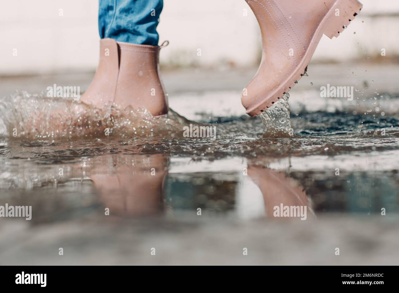 Frau mit Regengummistiefeln, die beim Herbstregen mit Wasserspritzern und Tropfen in die Pfütze laufen und springen Stockfoto