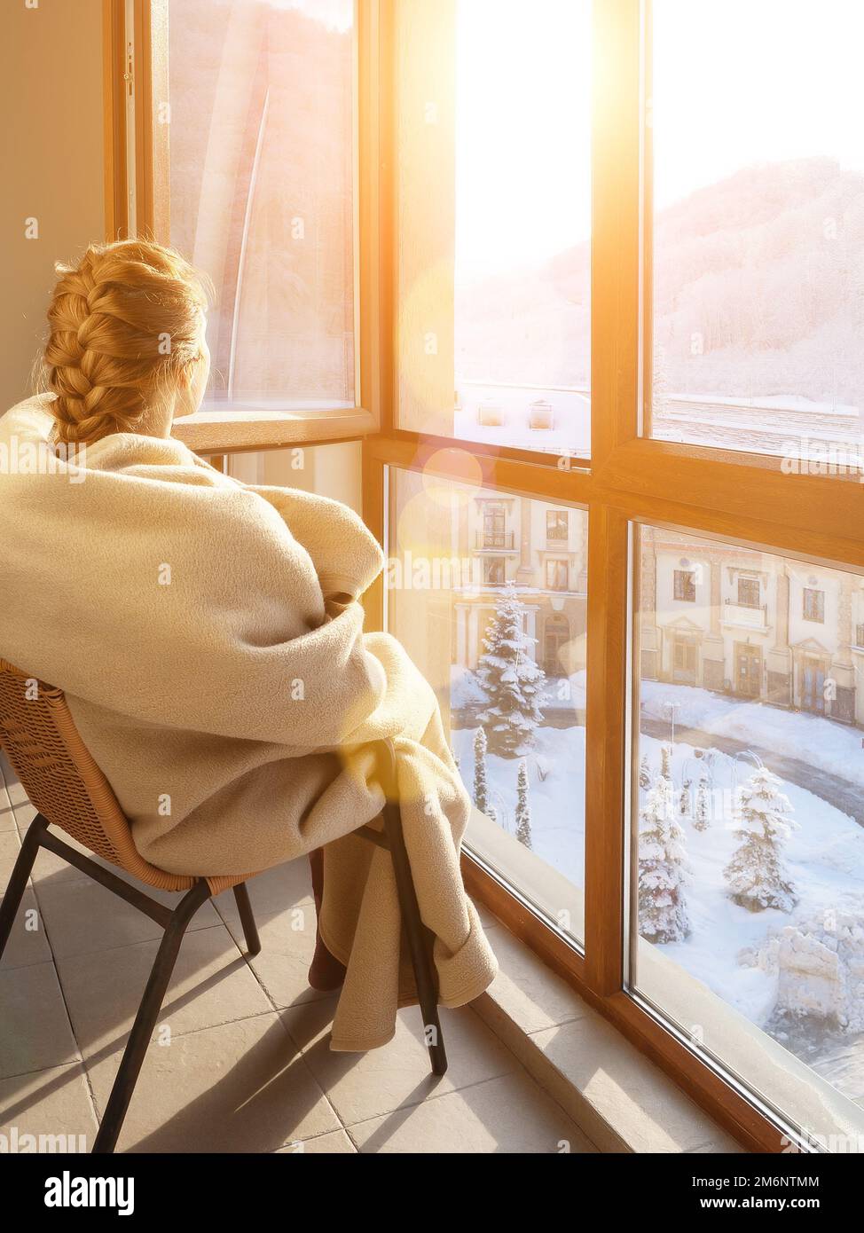 Eine in eine Decke gewickelte Frau sitzt an einem offenen Winterfenster. Eine Frau in einer Decke am Winterfenster. Ruhen Sie sich am Winterfenster aus Stockfoto