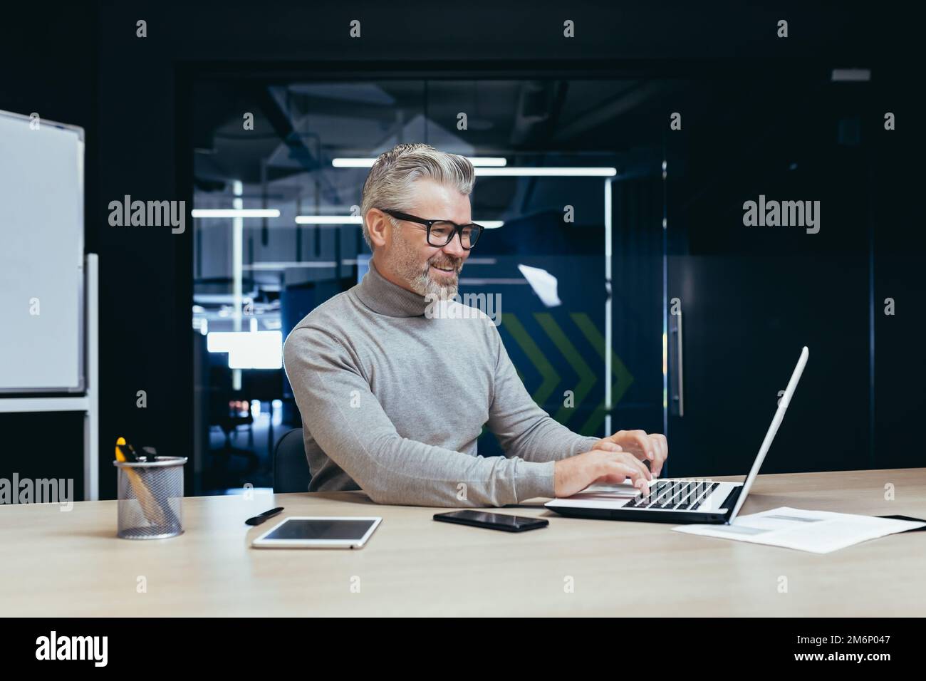 Fröhlicher und lächelnder, reifer Geschäftsmann, der im Büro arbeitet, älterer, grauhaariger Mann, der am Arbeitsplatz ein Notebook benutzt und mit guten Ergebnissen zufrieden lächelt. Stockfoto