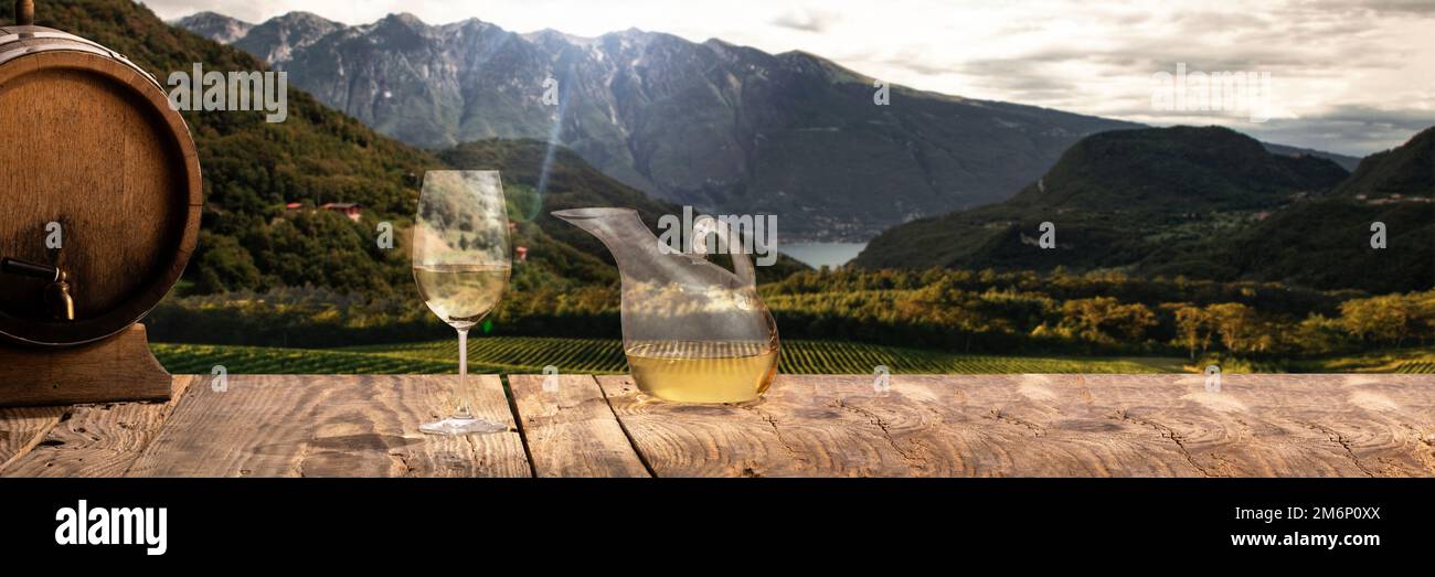 Karaffe, Glas mit Weißwein und Holzfass stehen auf einem Holztisch über einer wunderschönen Berg- und Feldlandschaft Stockfoto