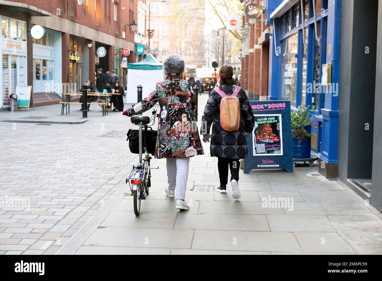 Rückansicht von zwei jungen Frauen, die entlang der Covent Garden Street Brompton Bike, wunderschöne bestickte Winterjacke Rucksack London UK 2022 KATHY DEWITT Stockfoto