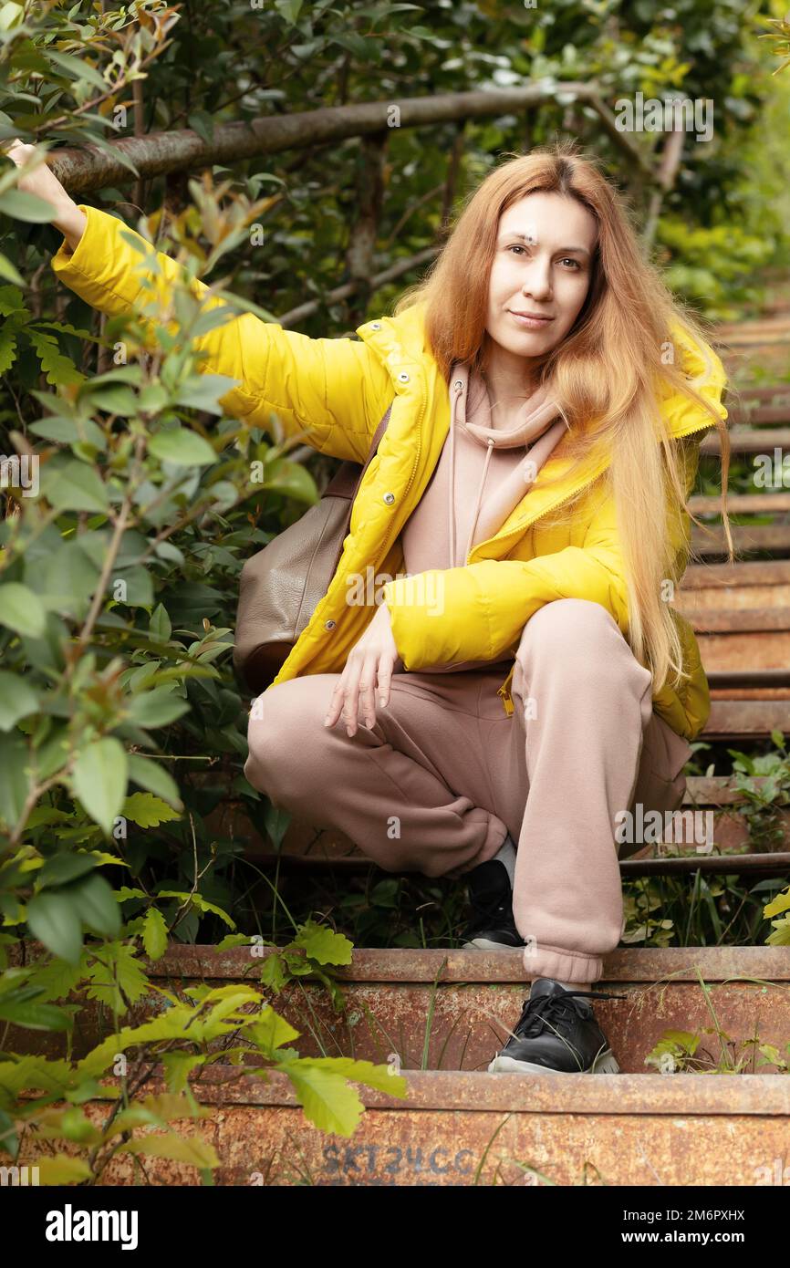 Eine blonde Frau in einer gelben Jacke sitzt auf der Treppe. Übergewachsene alte Treppen Stockfoto