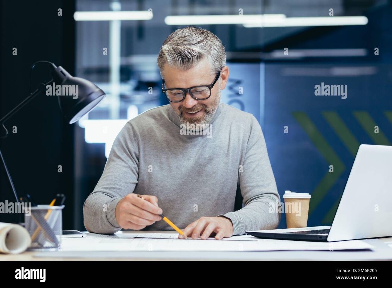 Erfolgreicher leitender Architekt bei der Arbeit im Büro, reifer, grauhaariger Mann, der Entwürfe zum Layout zeichnet und Pläne lächelt und mit der Arbeit zufrieden ist. Stockfoto