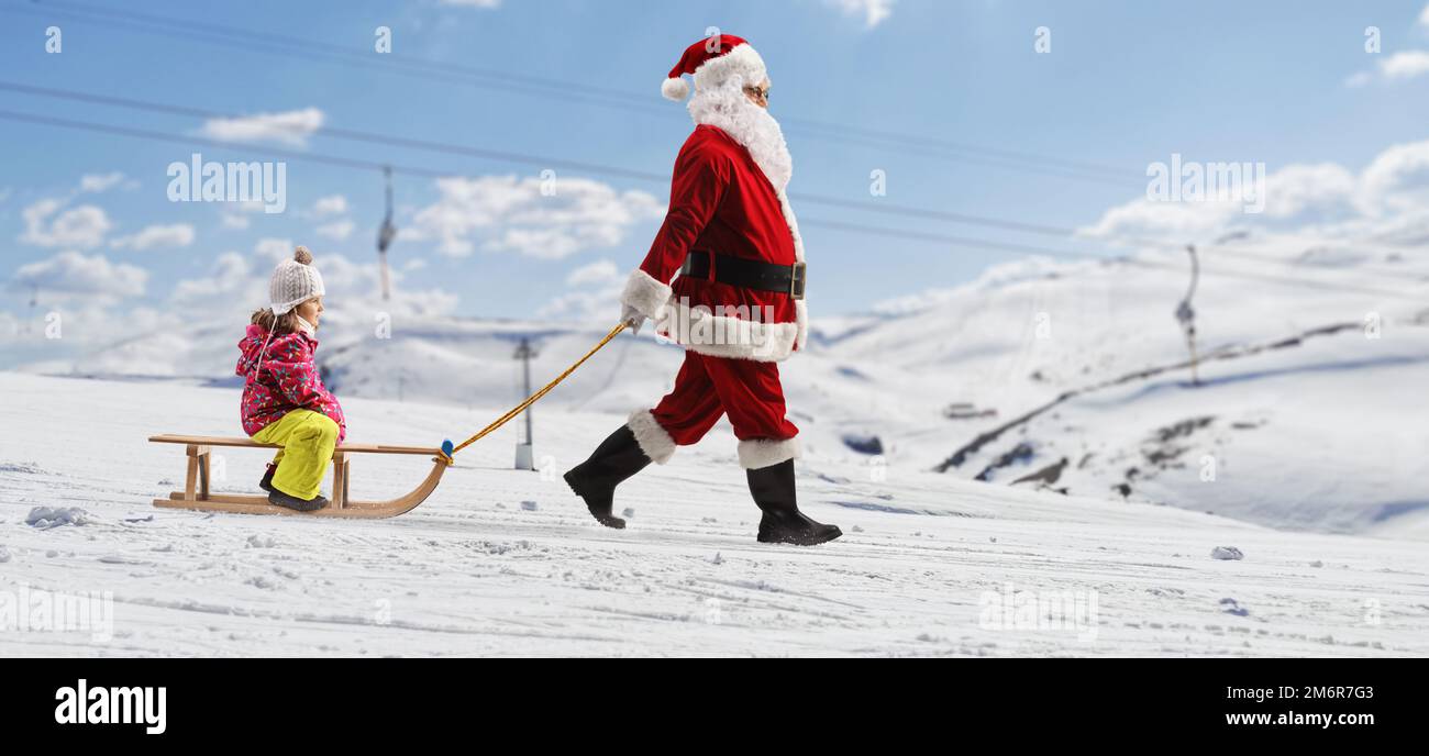 Profilaufnahme über die gesamte Länge, wie der weihnachtsmann ein Mädchen auf einem hölzernen Schlitten auf einem verschneiten Berg zieht Stockfoto