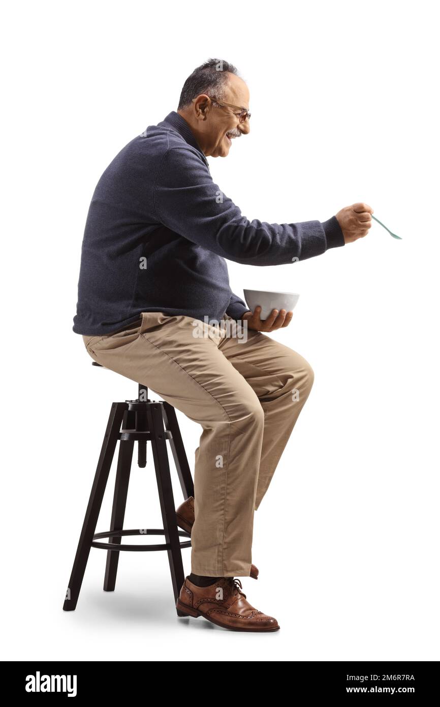 Profilaufnahme eines erwachsenen Mannes, der auf einem Stuhl sitzt und mit einem Löffel isst, isoliert auf weißem Hintergrund Stockfoto