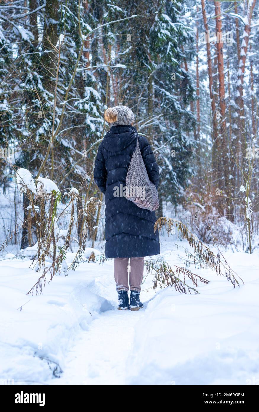 Eine Frau, die im Winterwald während eines Urlaubsspaziergangs zurücksteht, alleine im Wald mit einer Tragetasche, einem Strickmütze und einer Pufferjacke spaziert. Hochwertiges Foto Stockfoto