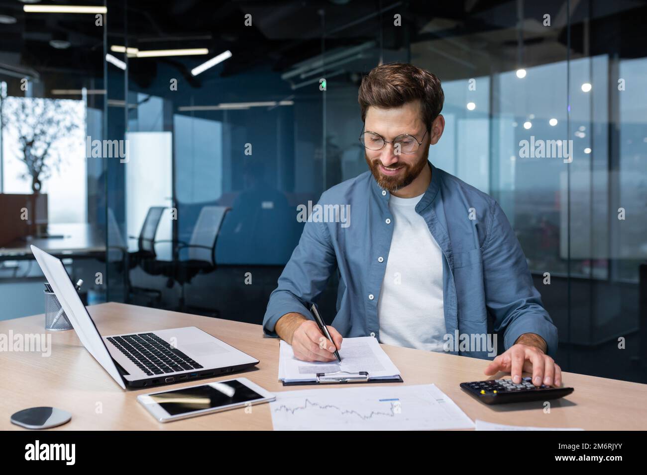 Erfolgreicher Finanzinvestor arbeitet mit Dokumenten und Konten im Büro, Geschäftsmann mit lächelndem Bart, zufrieden mit Arbeitsergebnissen und Erfolgen, Mann im Büro mit arbeitendem Laptop. Stockfoto