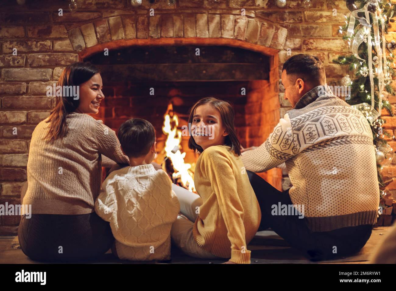 Glückliche junge Familieneltern mit zwei kleinen Kindern, die sich während der Weihnachtsferien am Kamin versammeln Stockfoto