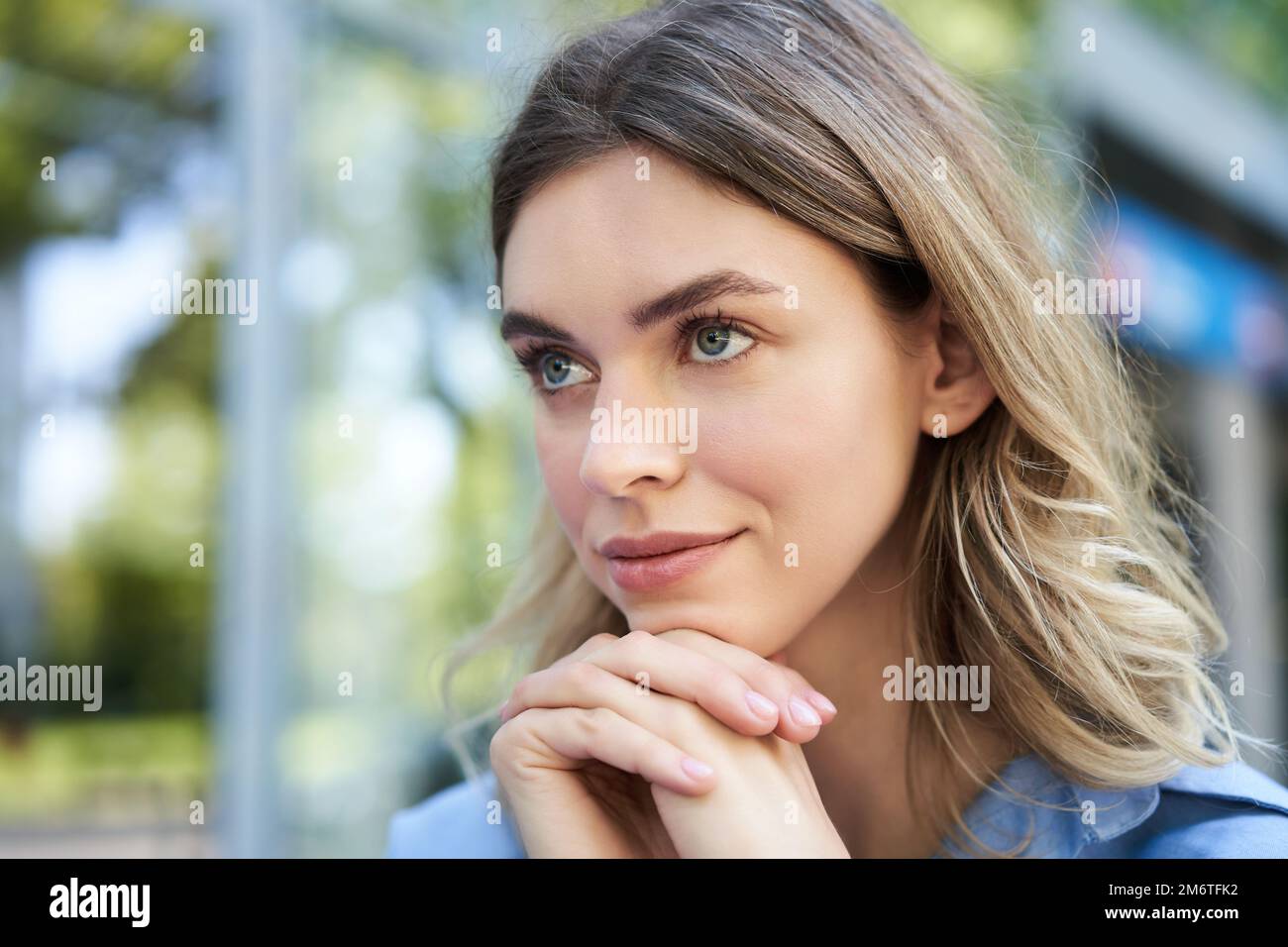 Nahaufnahme eines Porträts einer jungen Frau, die hoffnungsvoll und lächelnd aussieht, den Kopf auf die Hände lehnt, denkt, im Freien sitzt Stockfoto