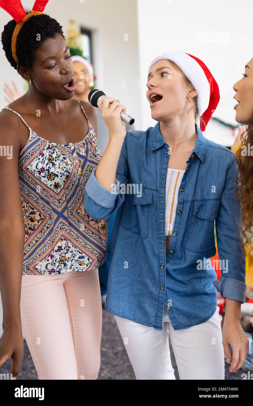 Vertikales Bild von fröhlichen, vielfältigen Freundinnen, die zu Hause weihnachten feiern und Karaoke singen Stockfoto