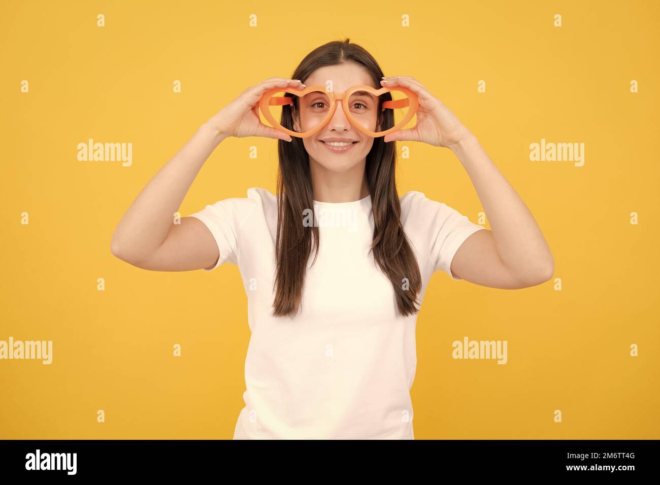 Portrait eines lustigen Mädchens mit coolen Partybrillen. Fröhliches junges Mädchen lächelt mit herzförmigen Gläsern auf gelbem Hintergrund. Stockfoto