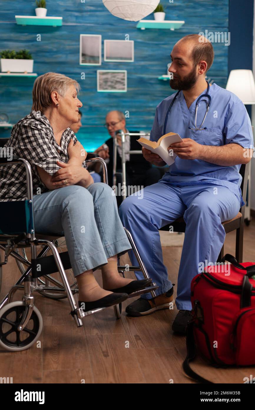 Männliche Krankenschwester, die Patienten durch Lesen von Geschichten aus Büchern an ältere behinderte Frauen im Wohnzimmer unterstützt. Pflegerische Sozialdienste für Senioren im Gesundheitszentrum. Stockfoto