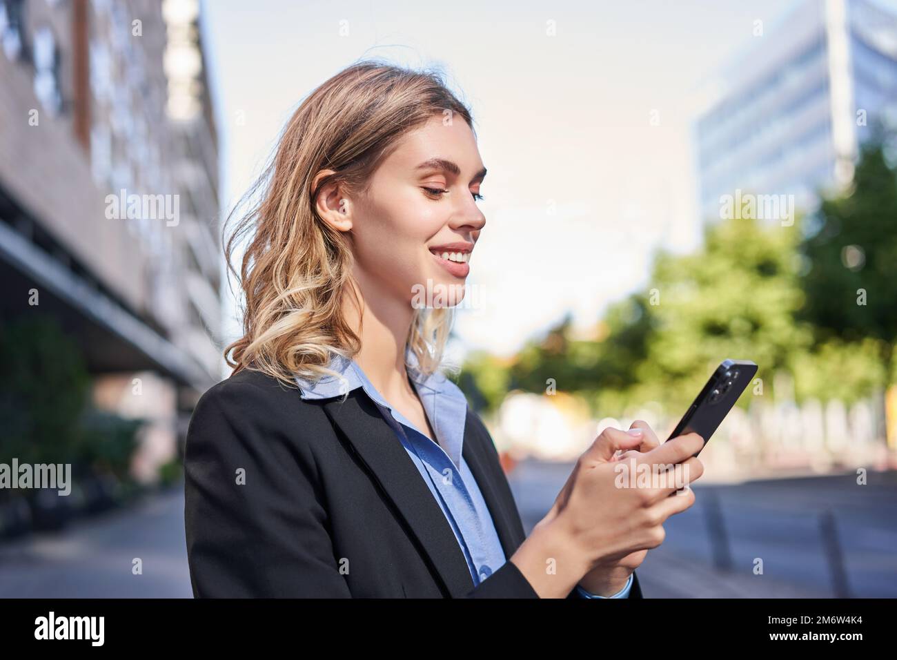 Eine Frau aus dem Unternehmen steht auf der Straße und SMS auf das Mobiltelefon, lächelt, während sie auf den Smartphone-Bildschirm schaut Stockfoto