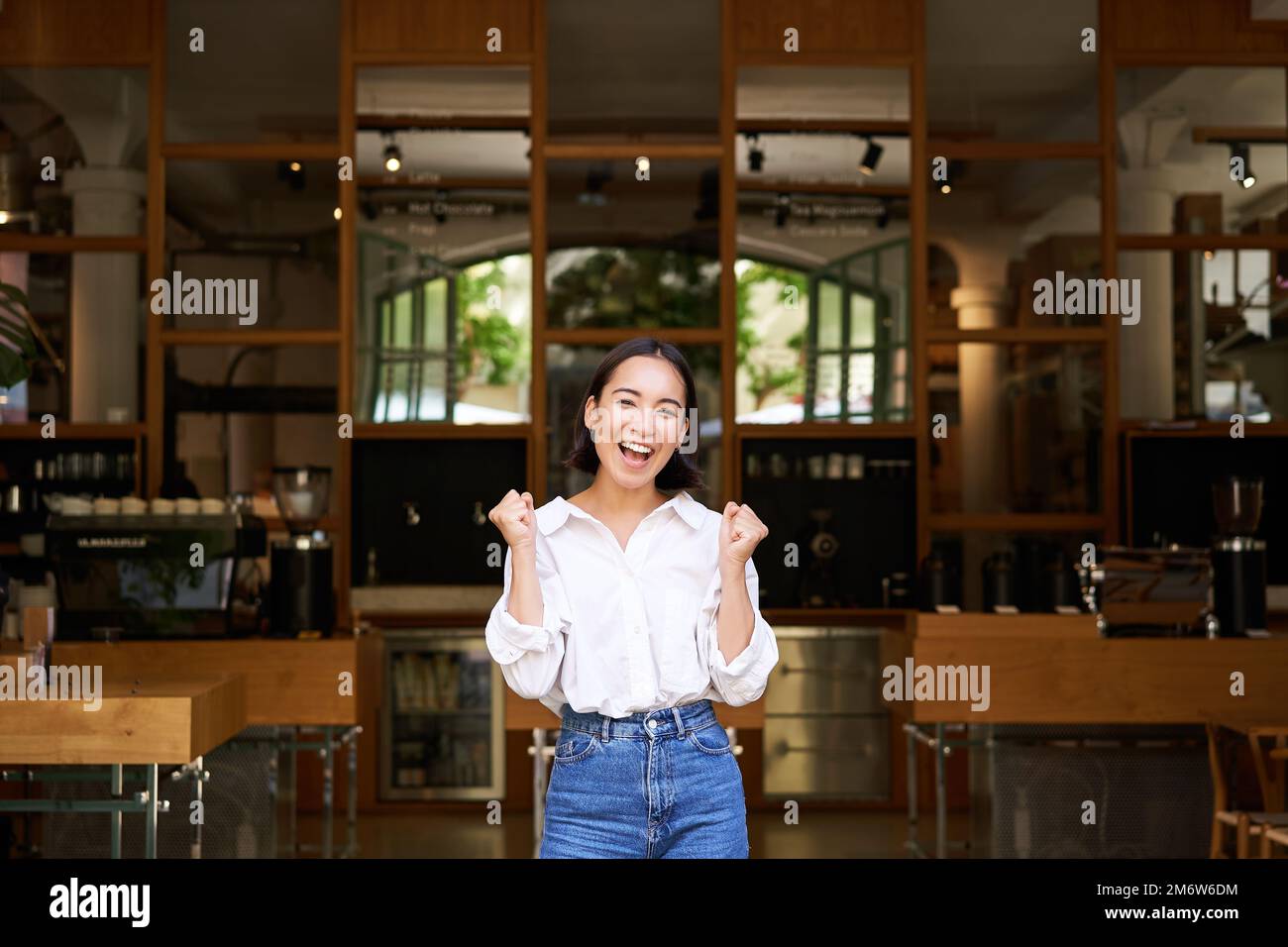 Glückliches, enthusiastisches asiatisches Mädchen, die Geschäftsbesitzerin triumphiert, eröffnet ihr eigenes Restaurant und steht vor einem leeren Café Stockfoto