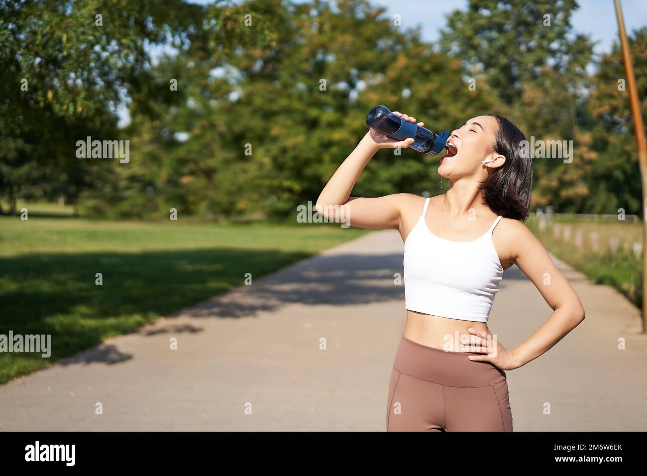 Glückliche asiatische Sportlerin, Läufer trinkt Wasser aus der Flasche beim Laufen, Training an der frischen Luft im Park Stockfoto
