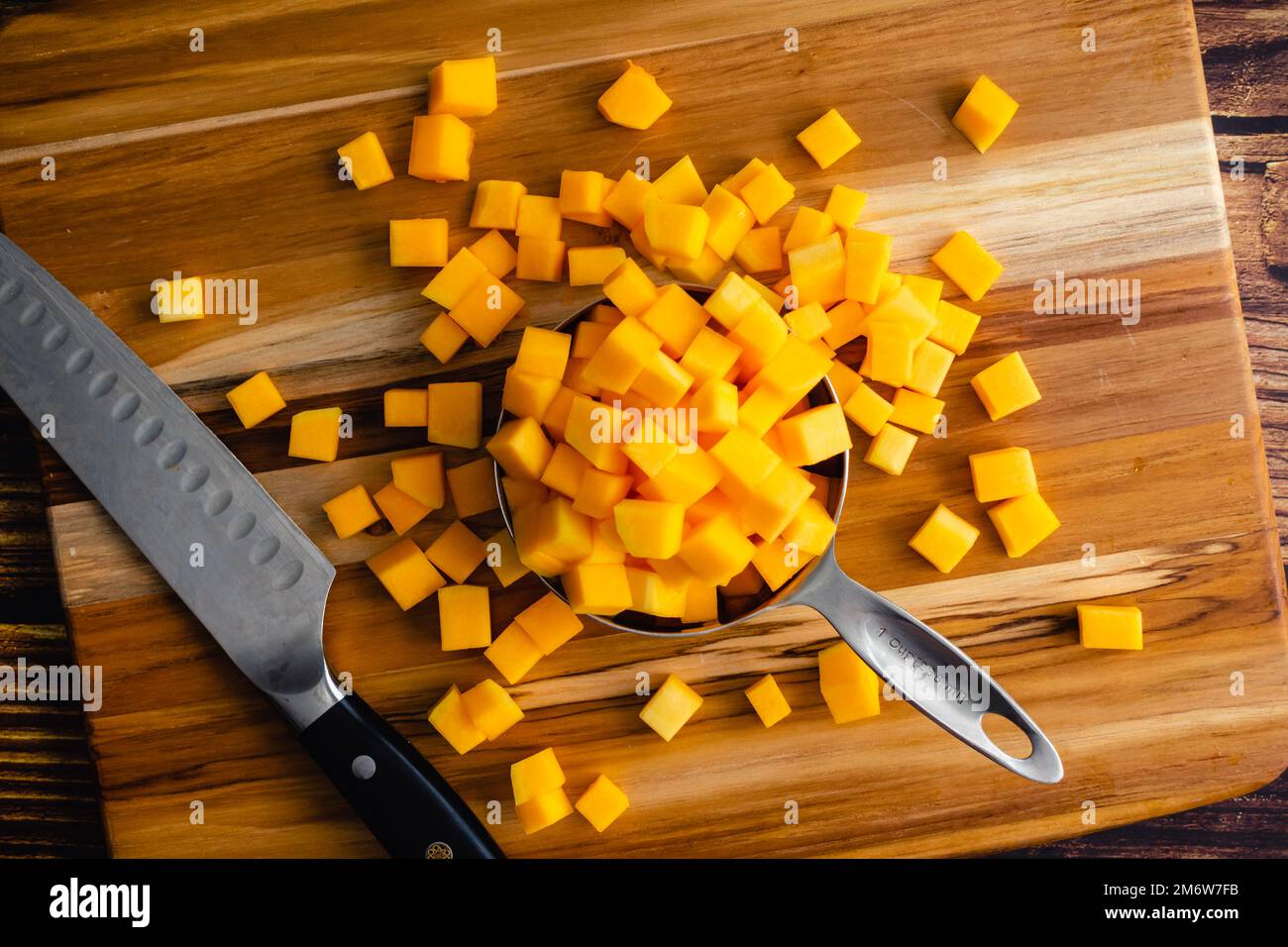 Butternusskürbis, der auf einem Holzschneidebrett in kleine Würfel geschnitten wurde: Gewürfelter Winterkürbis auf Holzhintergrund mit einem Küchenmesser Stockfoto