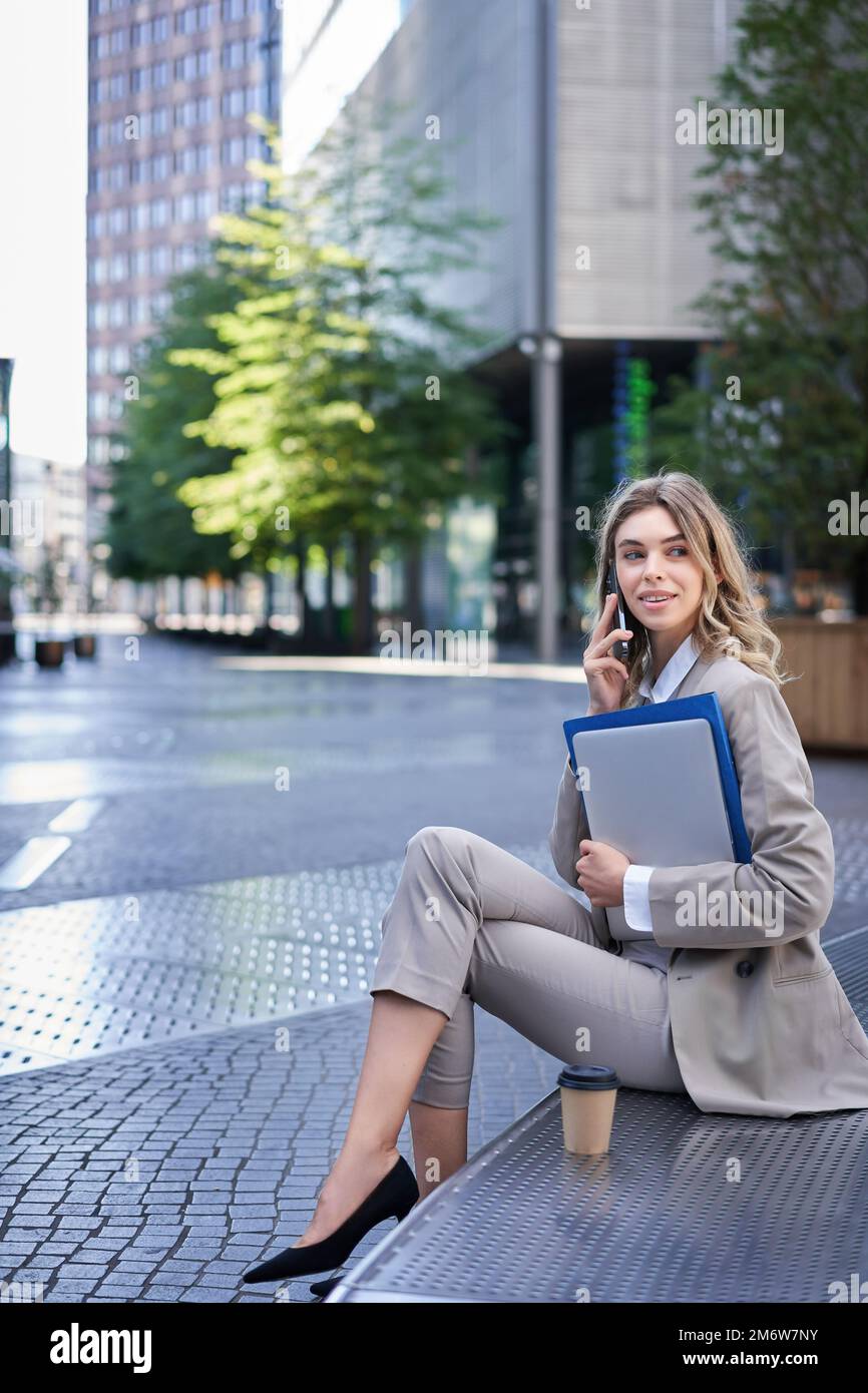 Vertikale Aufnahme einer jungen Frau, die in der Nähe eines Bürogebäudes sitzt, Laptop und Ordner mit Arbeitsdokumenten in der Hand hält, jemanden auf Mo anruft Stockfoto