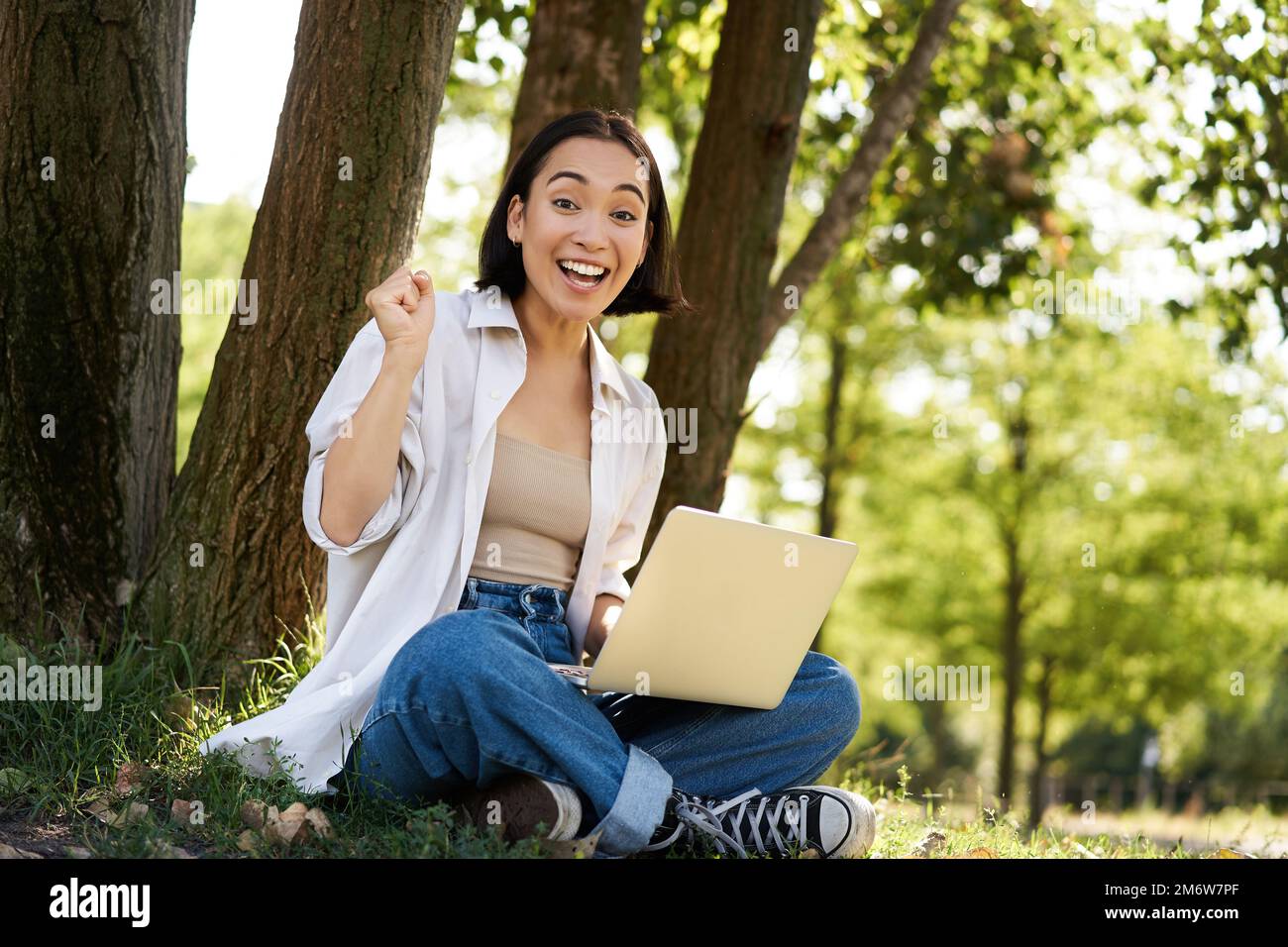 Enthusiastisches junges asiatisches Mädchen, das mit einem Laptop neben einem Baum im grünen, sonnigen Park sitzt, feiert, triumphiert und lächelt Stockfoto