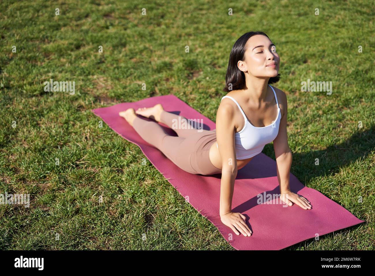 Lächelnde junge Sportlerin streckt sich im Park auf einer Gummimatte, macht Yoga-Asana-Übungen, trainiert an der frischen Luft in Fitnesskleidung Stockfoto