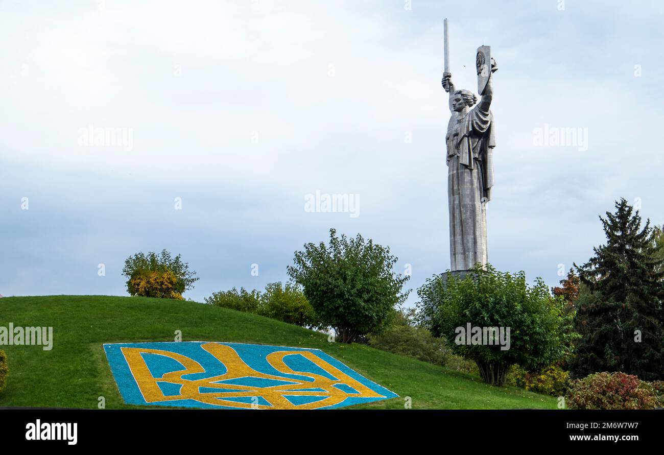 Die Statue des Vaterlands vor dem blauen Himmel. Ein Wappen der Ukraine auf dem Rasen, mit blauen und gelben Steinen gesäumt. Die Tride Stockfoto