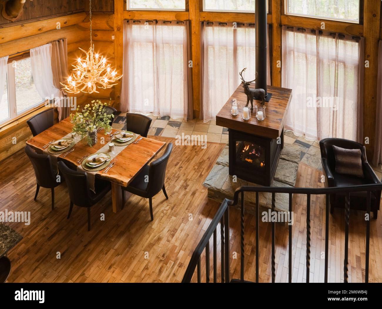 Blick aus dem Zwischengeschoss auf Esstisch aus Naturholz und braune Lederstühle im Esszimmer sowie Gaskamin im Wohnzimmer im luxuriösen Blockhaus. Stockfoto