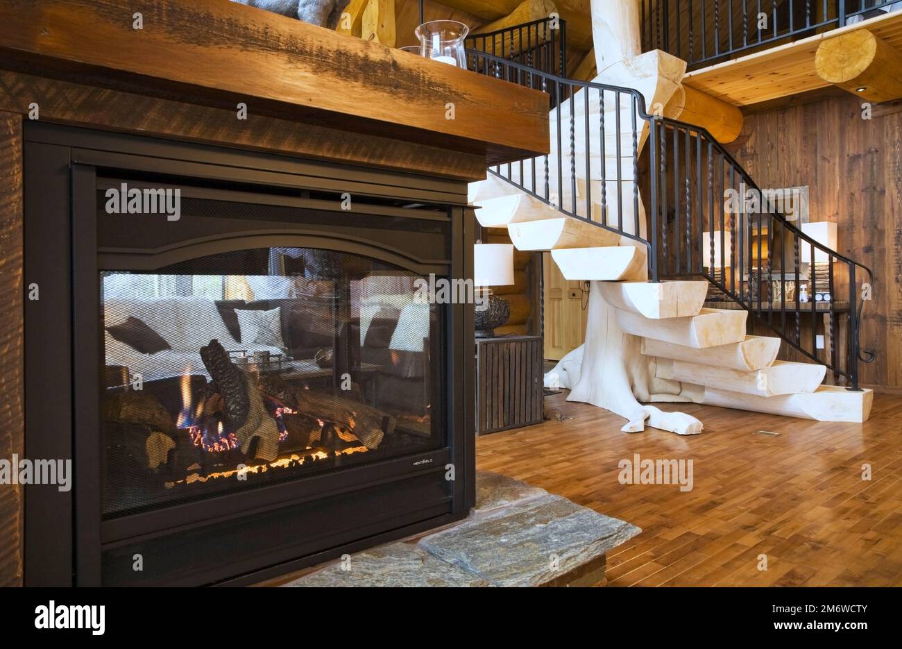 Gaskamin und gebleichte Holztreppen mit schwarzem schmiedeeisernem Geländer im Wohnzimmer im luxuriösen Blockhaus im Landhausstil. Stockfoto