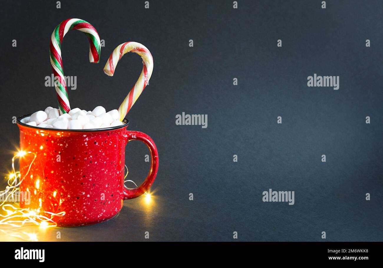 Roter Becher mit Marshmallows und Zuckerrohr in Herzform und Lichter von Girlanden auf schwarzem Hintergrund. Weihnachten Stockfoto