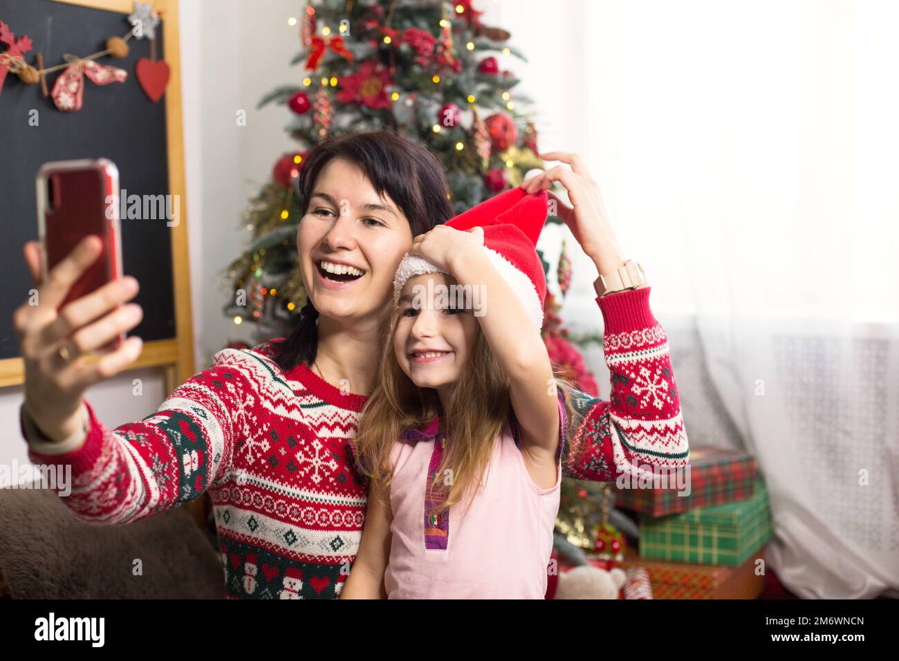 Mama und Tochter sitzen neben dem Weihnachtsbaum, umarmen sich, machen Fotos und Selfies am Telefon, kommunizieren über Video C. Stockfoto