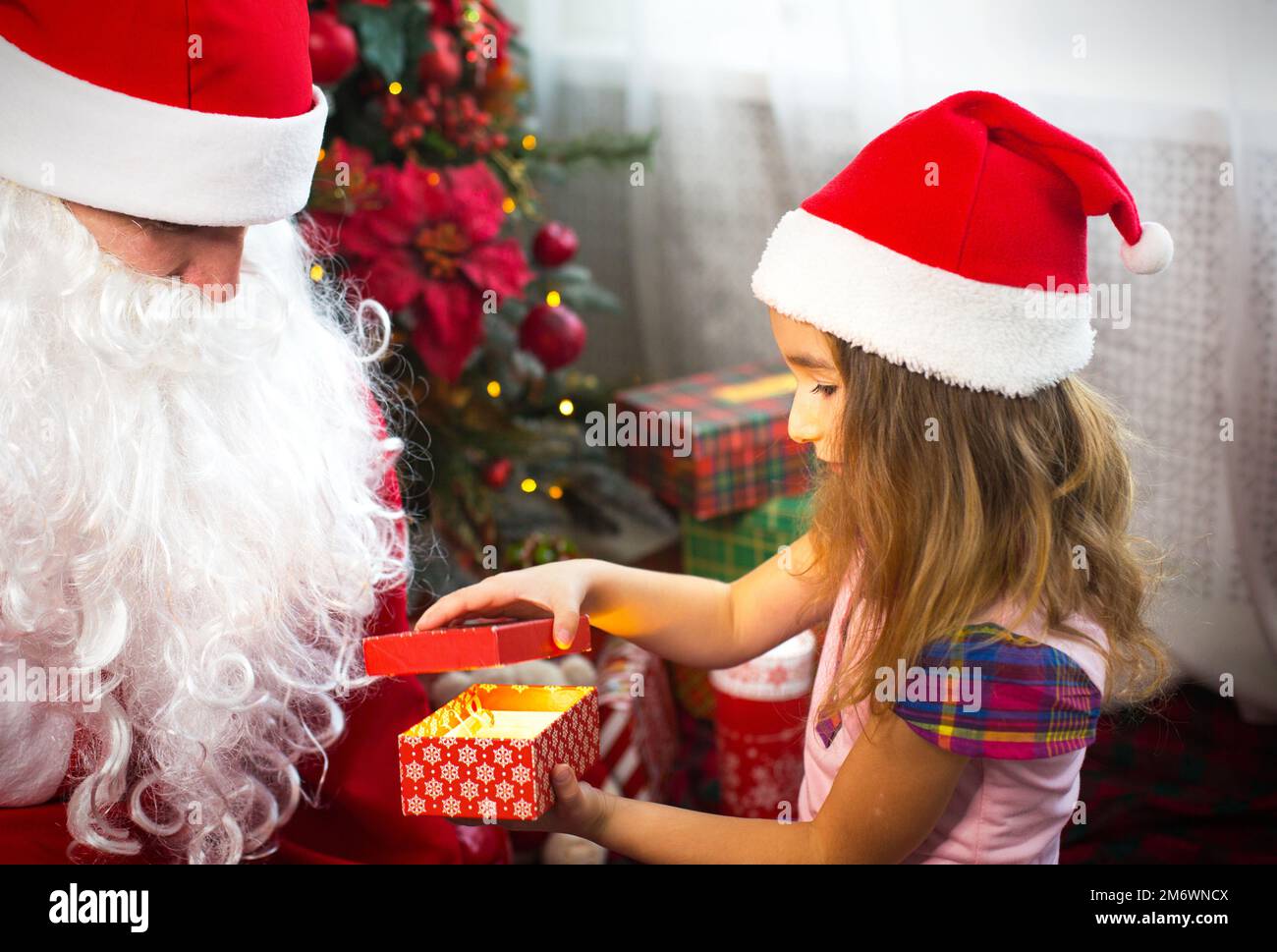 Ein kleines Mädchen mit Weihnachtsmannmütze auf dem Schoß des Weihnachtsmanns neben dem Weihnachtsbaum im Weihnachtsdekor. Zeigt eine Schachtel mit einem Geschenk, ein weiches Spielzeug, das seine berührt Stockfoto