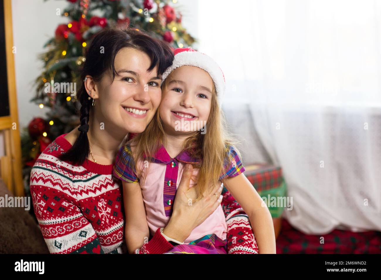 Mama und Tochter umarmen sich, lächeln fröhlich und schauen in die Kamera, sitzen zu Hause am Weihnachtsbaum in bequemen Kleidern und einem San Stockfoto