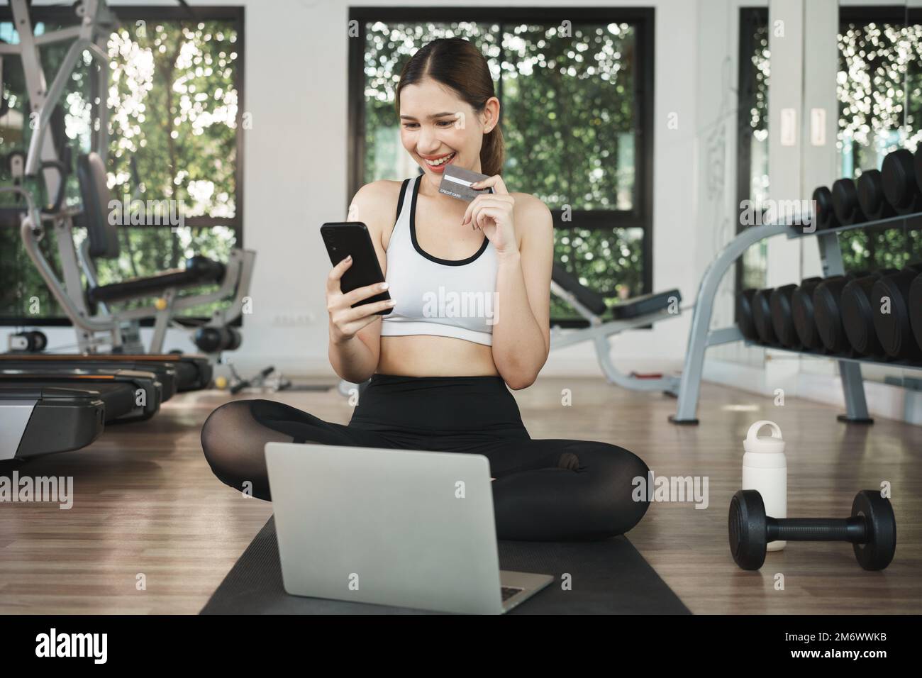 Glückliche Frau, die Handy und Kreditkarte benutzt, um im Fitnessstudio online einzukaufen. Stockfoto