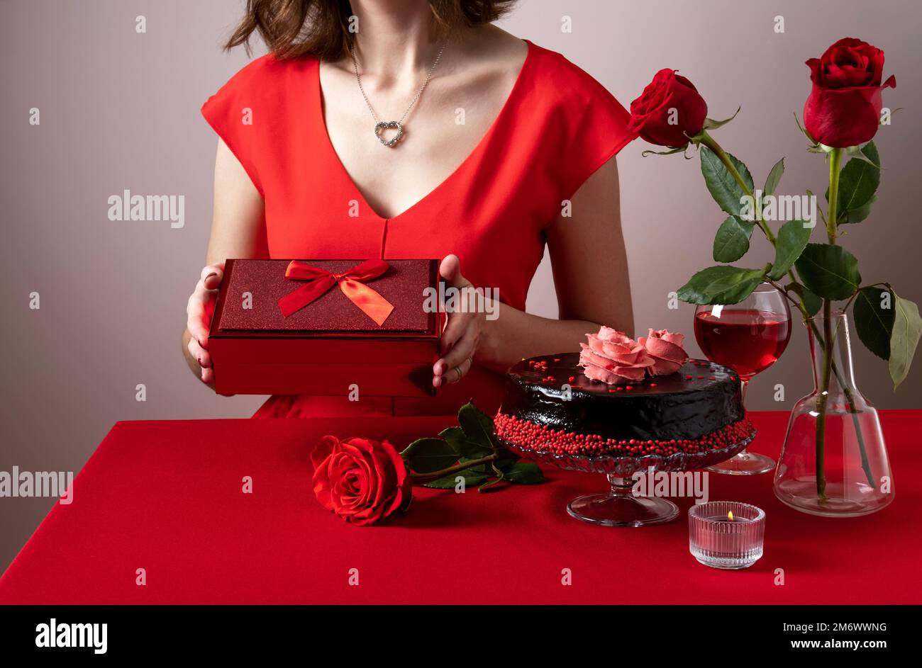Eine Frau in rotem Kleid, die eine rote Geschenkbox in der Hand hält. Abendessen am Valentinstag mit Tischdecken, Rotwein, Kerzen und Schokolade Stockfoto