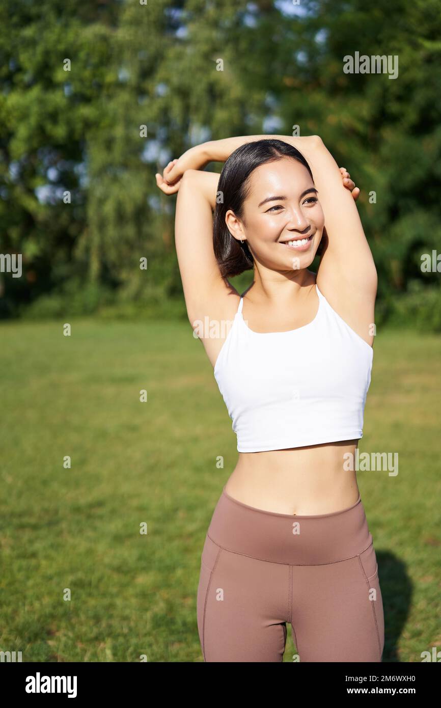 Asiatische Frau streckt sich die Arme, macht ein Fitnesstraining im Park, lächelt zufrieden, wärmt sich auf, bevor sie tagsüber an der frischen Luft joggt Stockfoto
