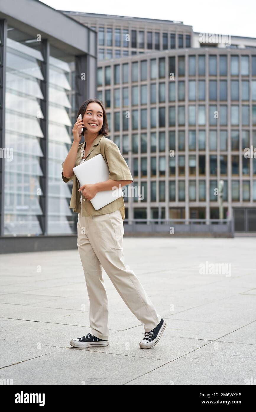 Vertikales Porträt einer stilvollen asiatischen Schülerin, die beim Gehen mit dem Handy spricht und ein Notebook in der Hand hält Stockfoto