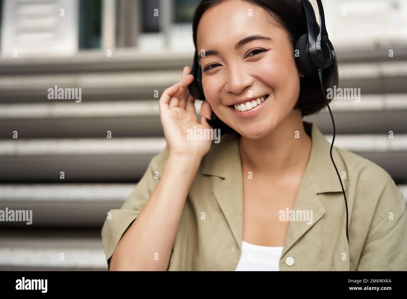 Glückliches asiatisches Mädchen hört Musik über Kopfhörer, lächelt, sitzt draußen auf der Treppe Stockfoto