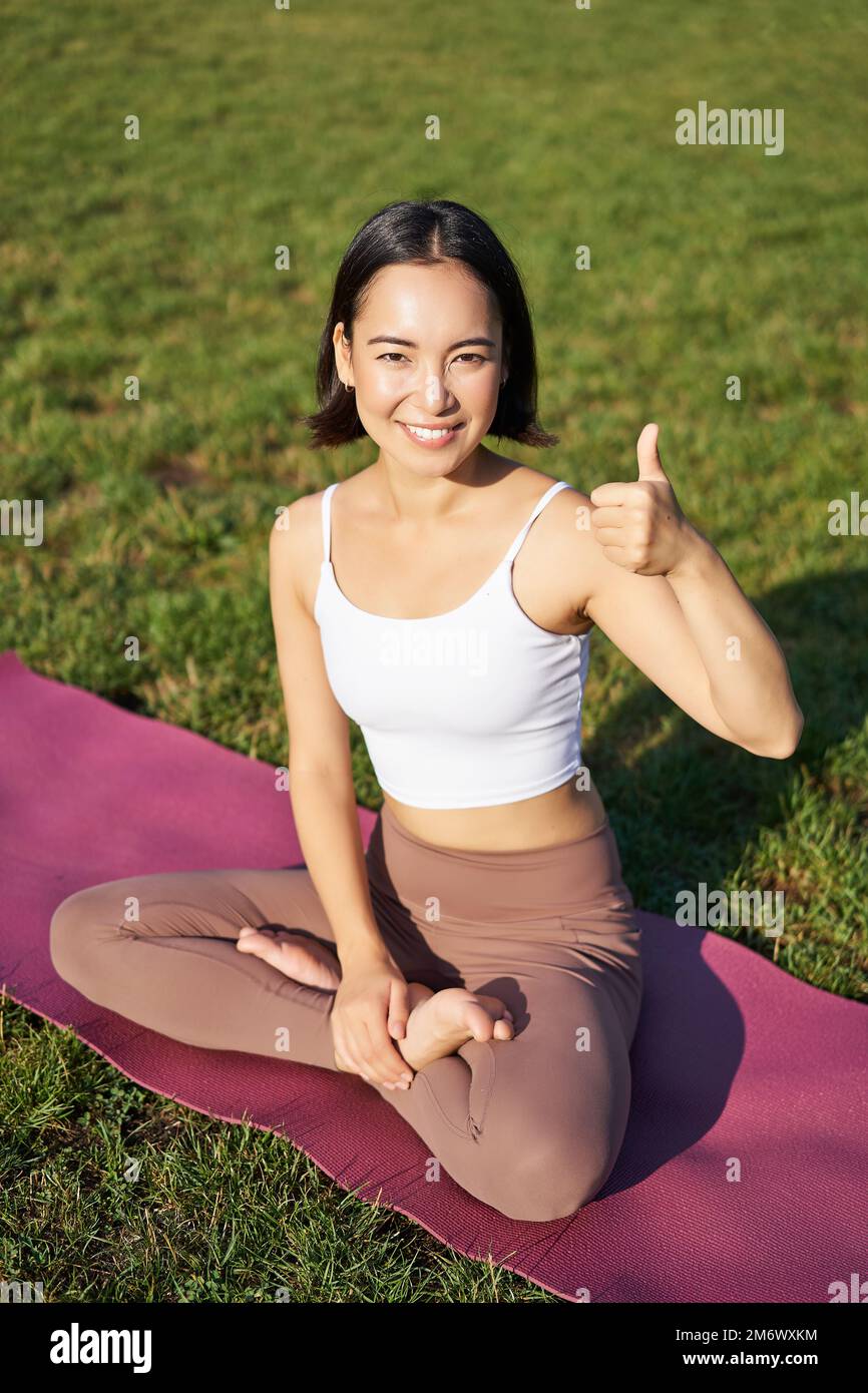 Vertikale Aufnahme einer asiatischen Frau, die auf einer Gummimatte sitzt, nach einem guten Yoga-Training im Park, mit "Daumen nach oben" als Zustimmung. Ich meditiere weiter Stockfoto