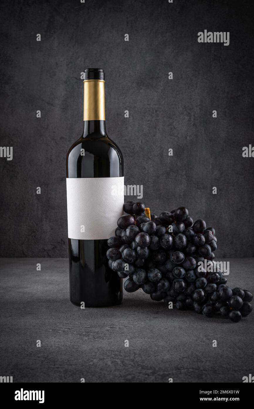 Flasche Rotwein mit Trauben auf dunklem Hintergrund. Rotwein mit Früchten. Weinflaschenmodell mit leeren Etiketten zum Anbringen Stockfoto