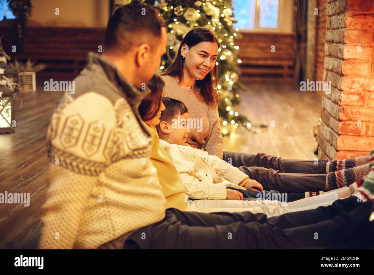 Glückliche junge Familieneltern mit zwei kleinen Kindern, die sich zu Weihnachten um den Ziegelkamin in einem gemütlichen und warmen Landhaus versammeln Stockfoto