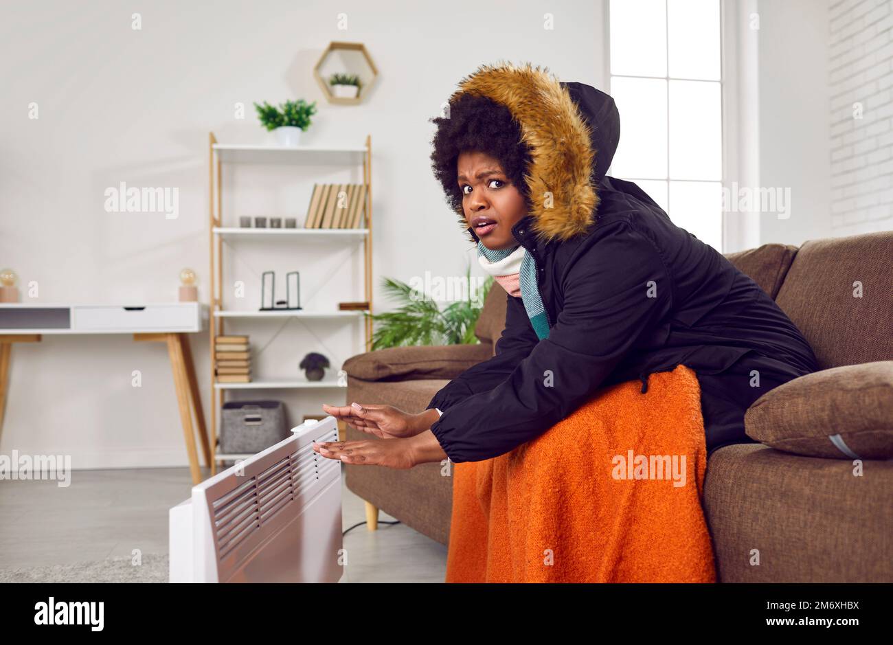 Eine afroamerikanische Frau im Wintermantel auf dem Sofa und wärmt sich die Hände mit einem elektrischen Heizgerät Stockfoto