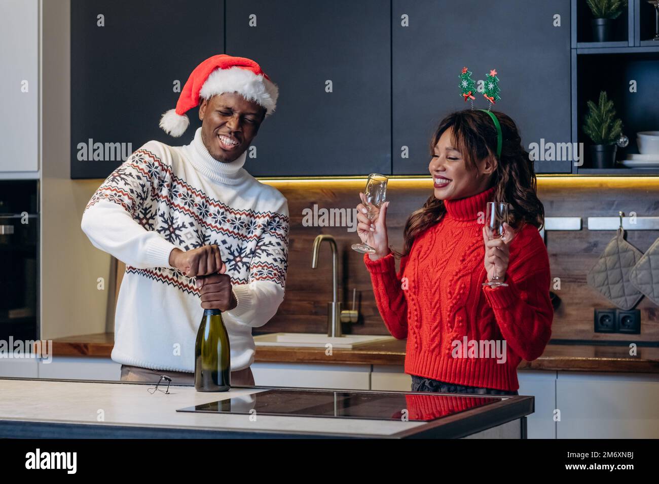 Eine junge weiße Frau und ein schwarzer Mann mit festlichen Hüten und bunten bedruckten Pullover schütten Champagner in die Weingläser, um Weihnachten zu feiern Stockfoto