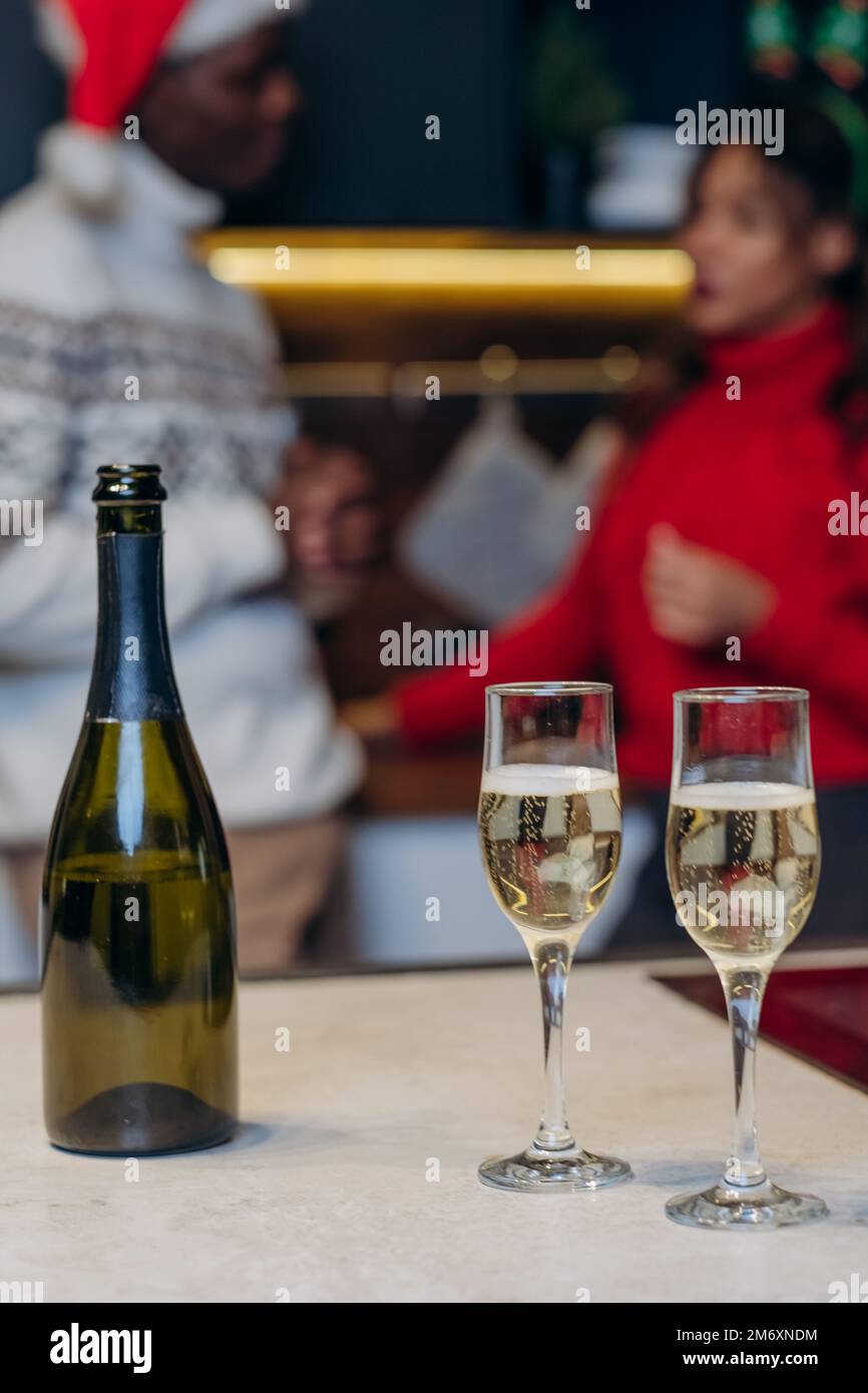 Weingläser mit Champagner und Flasche auf dem Tisch gegen ein fröhliches Paar in festlichen Strickpullover, das in weihnachtlicher Atmosphäre kommuniziert Stockfoto