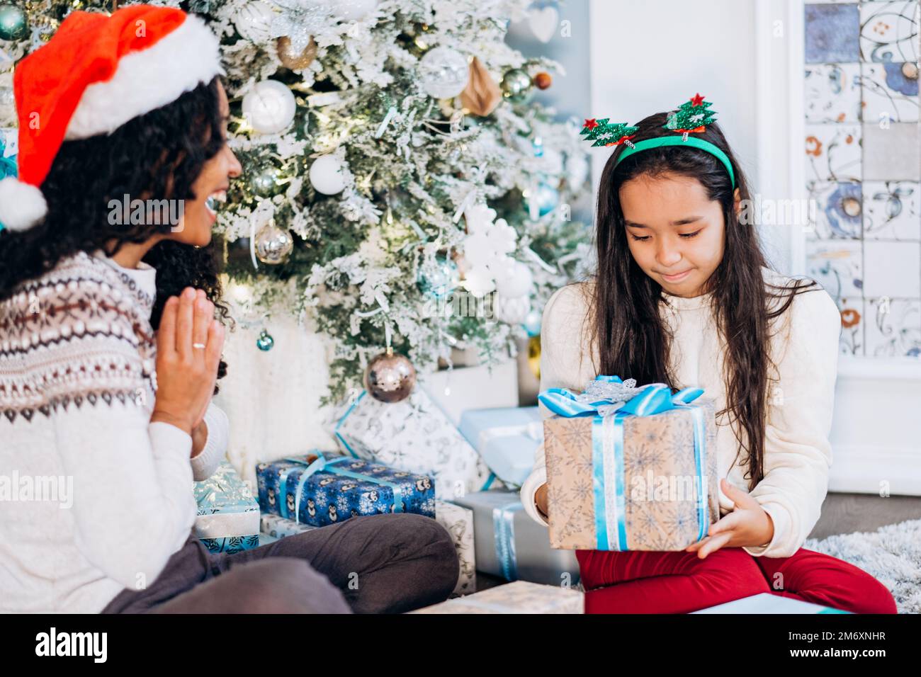 Lockige, langhaarige afroamerikanische Mutter und brünette Tochter sitzen auf dem Teppich und öffnen festliche Geschenke, die in der Nähe des Weihnachtsbaums fröhlich lächeln Stockfoto