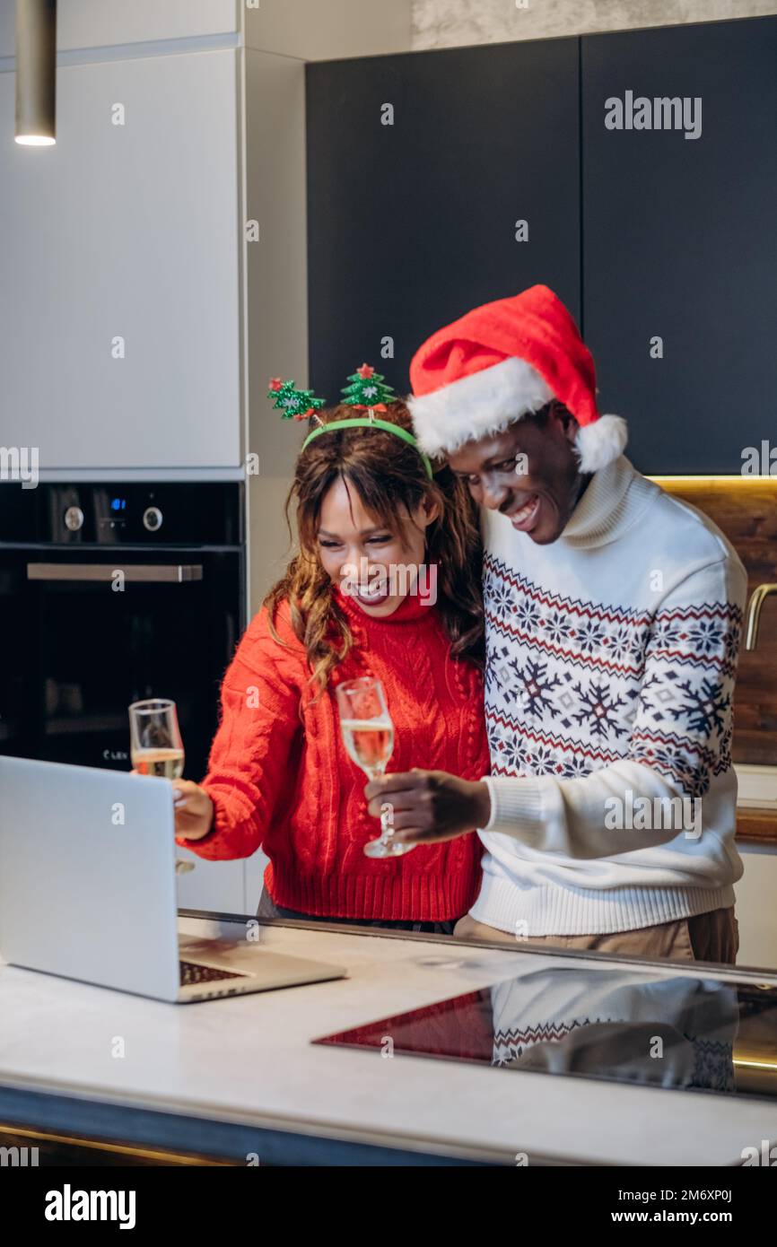 Junge Frau in festlichem Pullover und schwarzer Mann mit lustigem Hut sprechen per Videoanruf über Laptop und feiern Weihnachten in der Küche Stockfoto
