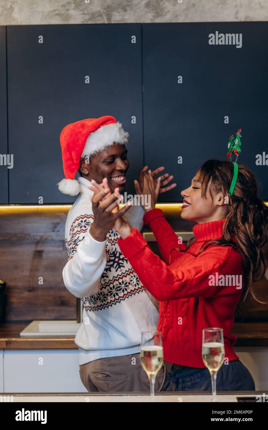 Ein afroamerikanischer Mann mit Weihnachtsmannmütze und eine lächelnde weiße Frau im Pullover tanzen in weihnachtlicher Atmosphäre und trinken Champagner in der Küche Stockfoto