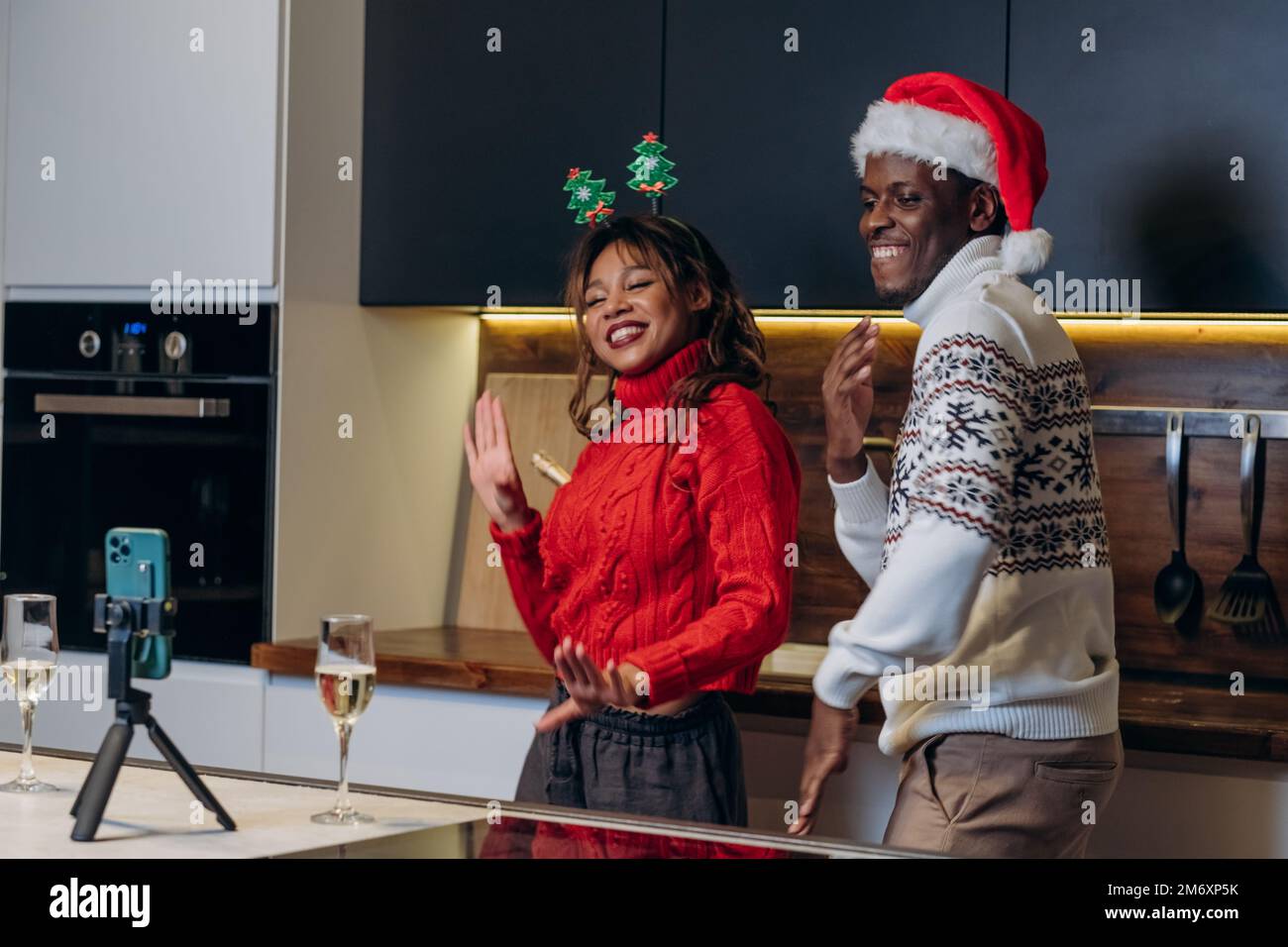 Ein afroamerikanischer Mann mit Weihnachtsmannmütze und eine lockige Freundin in warmen Pullover tanzen in Weihnachtsstimmung und Filmen ein Video mit einem Smartphone auf einem Stativ in der Küche Stockfoto
