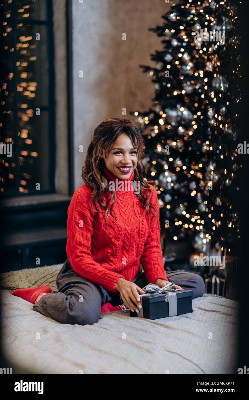 Junge Frau mit lockigem Haar in festlichem, rotem Strickpullover lächelt mit einem Geschenk und posiert gegen den Weihnachtsbaum Stockfoto
